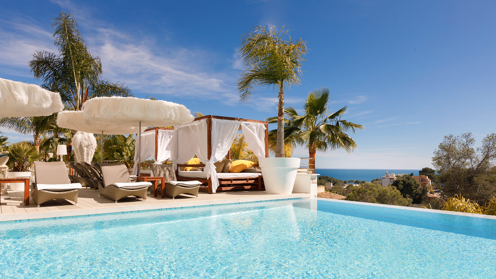 A view from the pool, with sun loungers, parasols, daybeds and palm trees, of the Mediterranean Sea on the horizon.